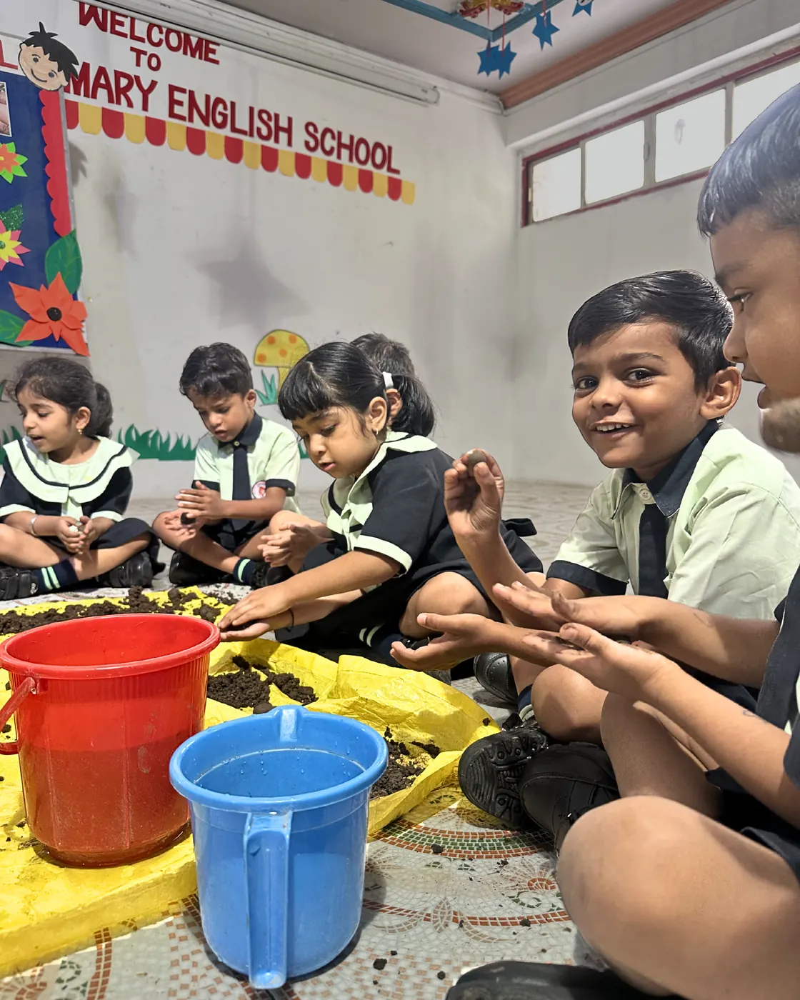 Children with buckets of soil and seeds on the classroom floor.